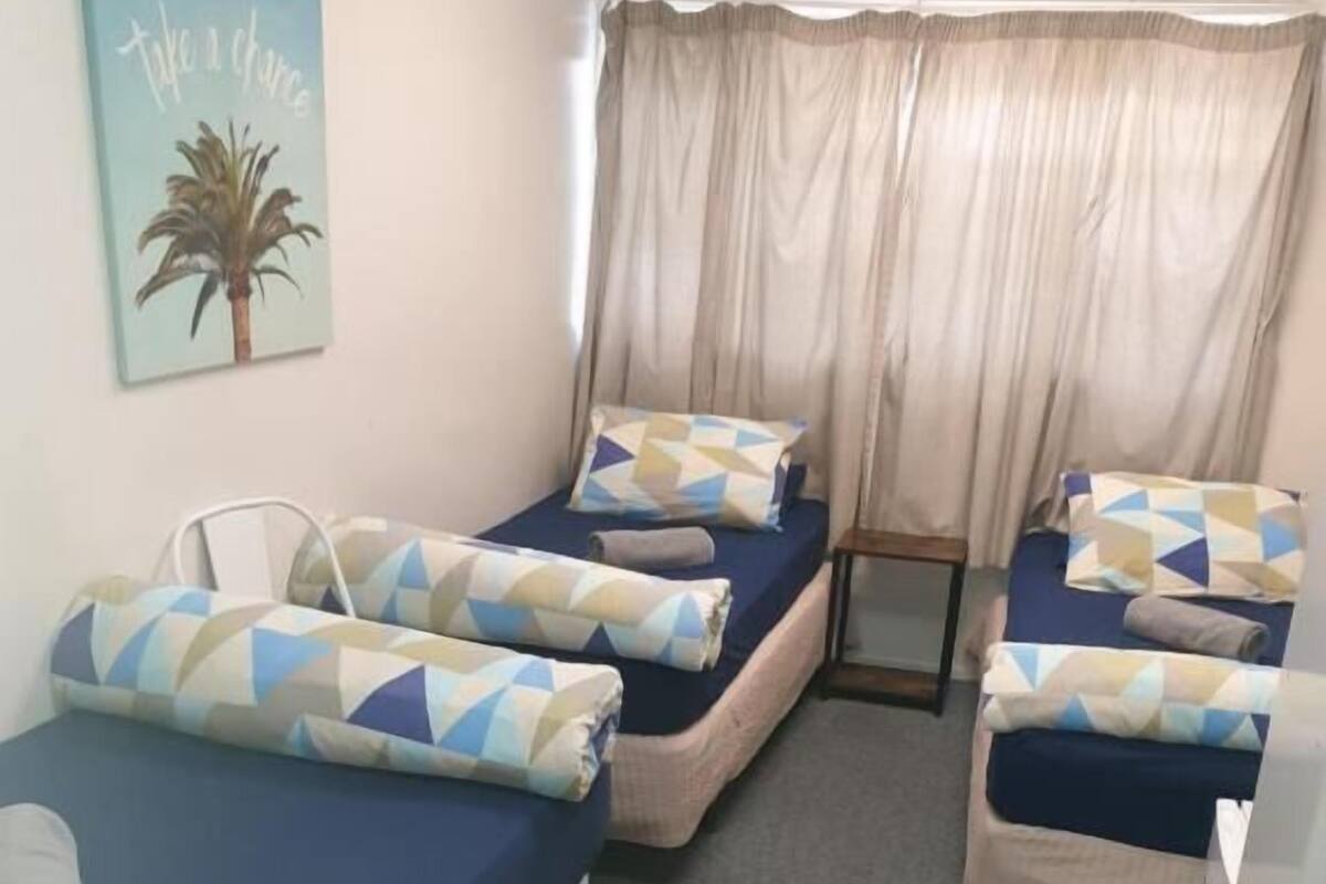 Quarto com três camas de solteiro, cada uma com colchão azul e almofadas estampadas. Há uma pequena mesa e uma cadeira no centro, uma janela com cortinas e um quadro pendurado na parede.