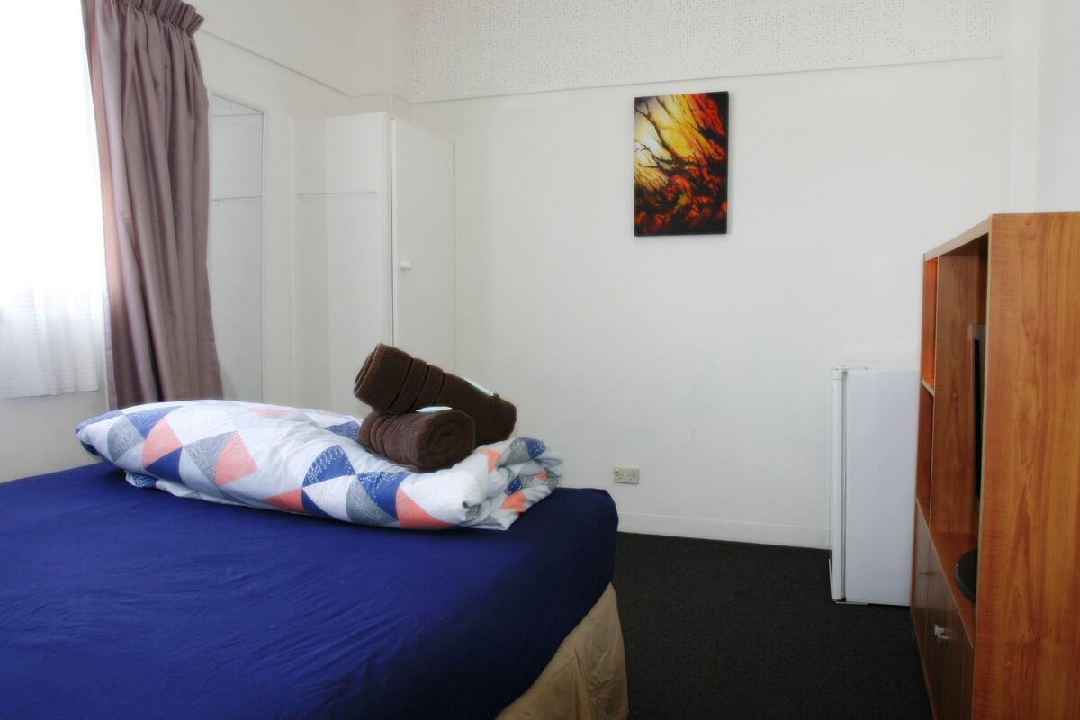 Uma cama de solteiro com colcha azul e almofada estampada em padrão geométrico, manta marrom e travesseiro branco. Há uma parede branca com um quadro abstrato colorido e uma porta branca. Um armário de madeira é visível à direita.