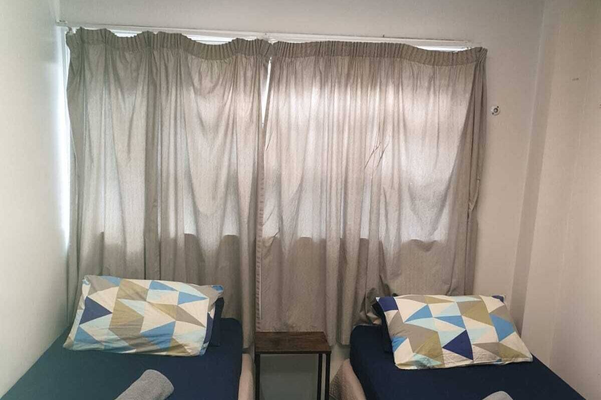 Acomodação pequena, com um quarto e duas camas de solteiro, cada uma com roupa de cama estampada e uma toalha enrolada. Uma pequena mesa está colocada entre as camas.