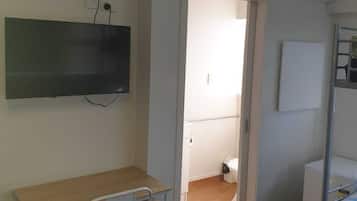 Ein schlichter, zweckmäßiger Raum mit Schreibtisch, Stuhl und Bett. Ein Fernseher ist an der Wand befestigt, es gibt eine Klimaanlage und eine Tür zu einem weiteren Raum.