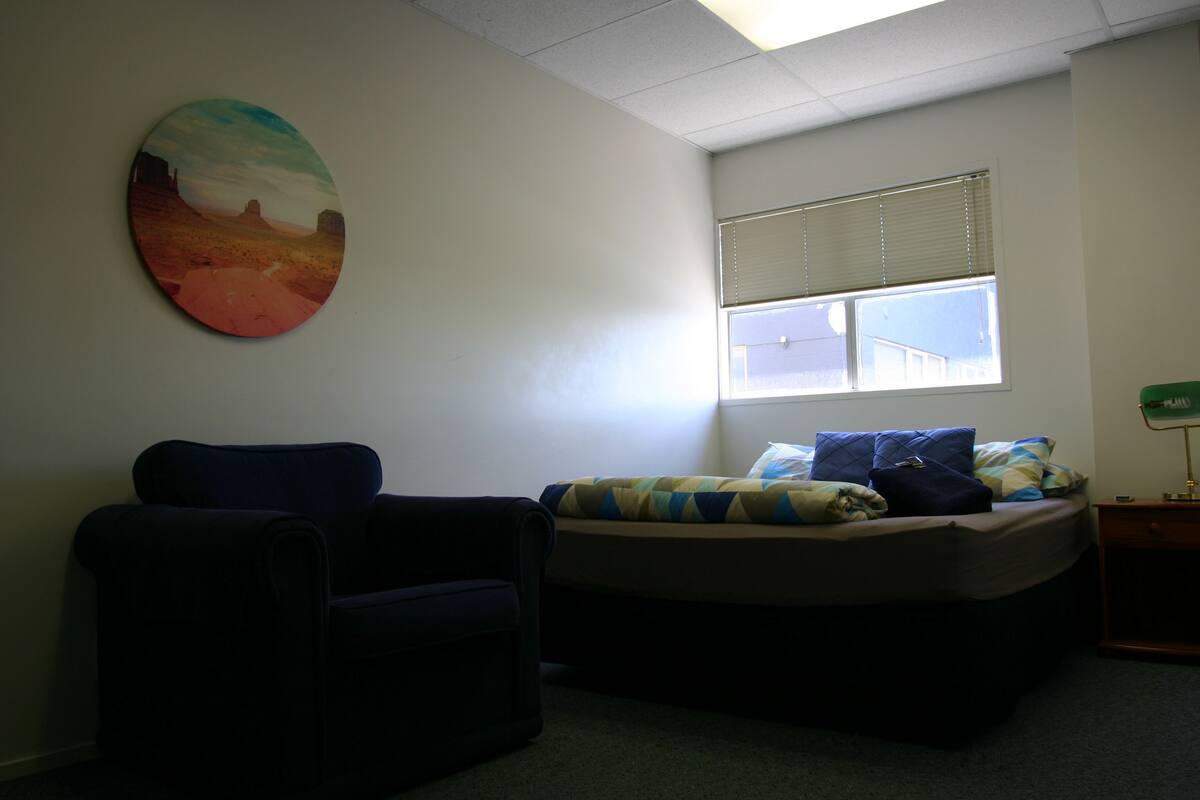 Quarto com cama, cadeira, janela com persianas e um quadro na parede.