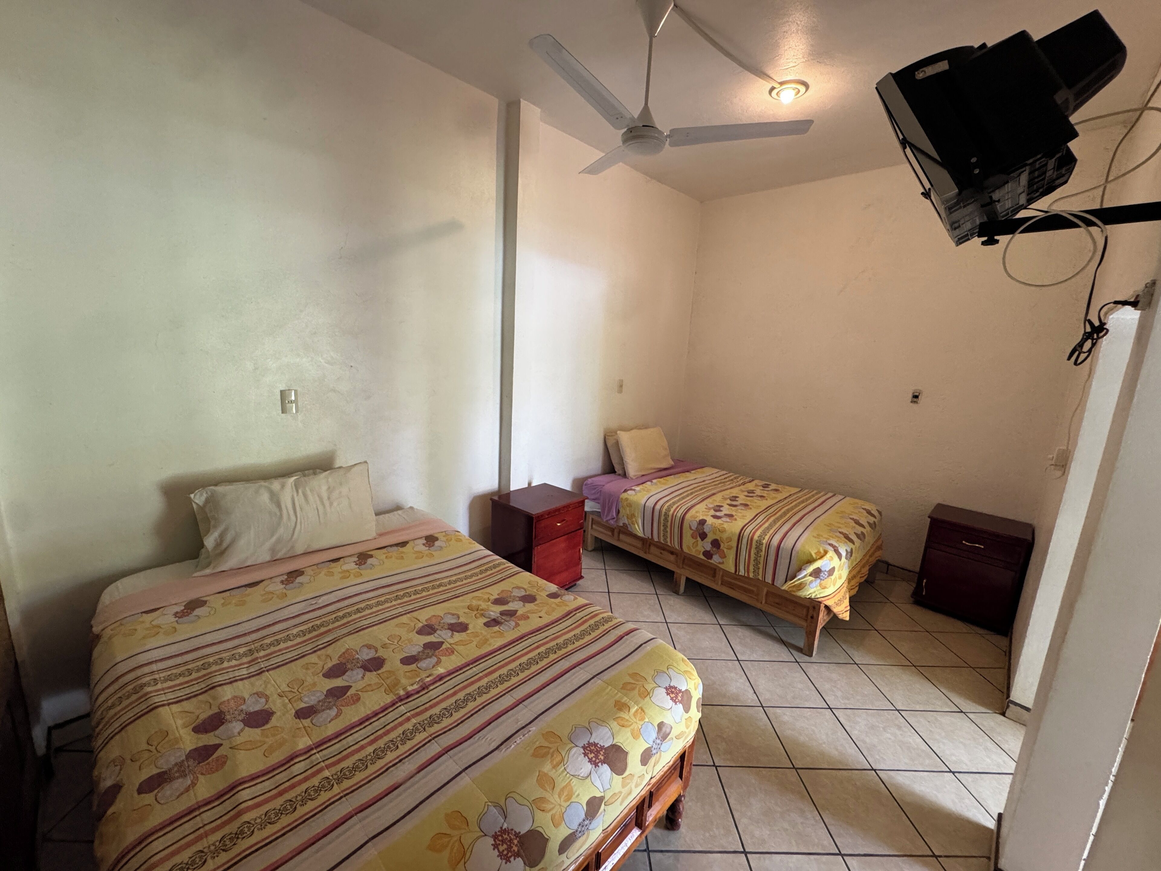A small, well-lit bedroom with two single beds, a ceiling fan, and a projector mounted on the wall.