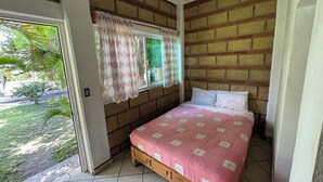 Un dormitorio con pared de ladrillo, ventana con cortinas florales, cama con colcha rosa y vista a follaje exterior.
