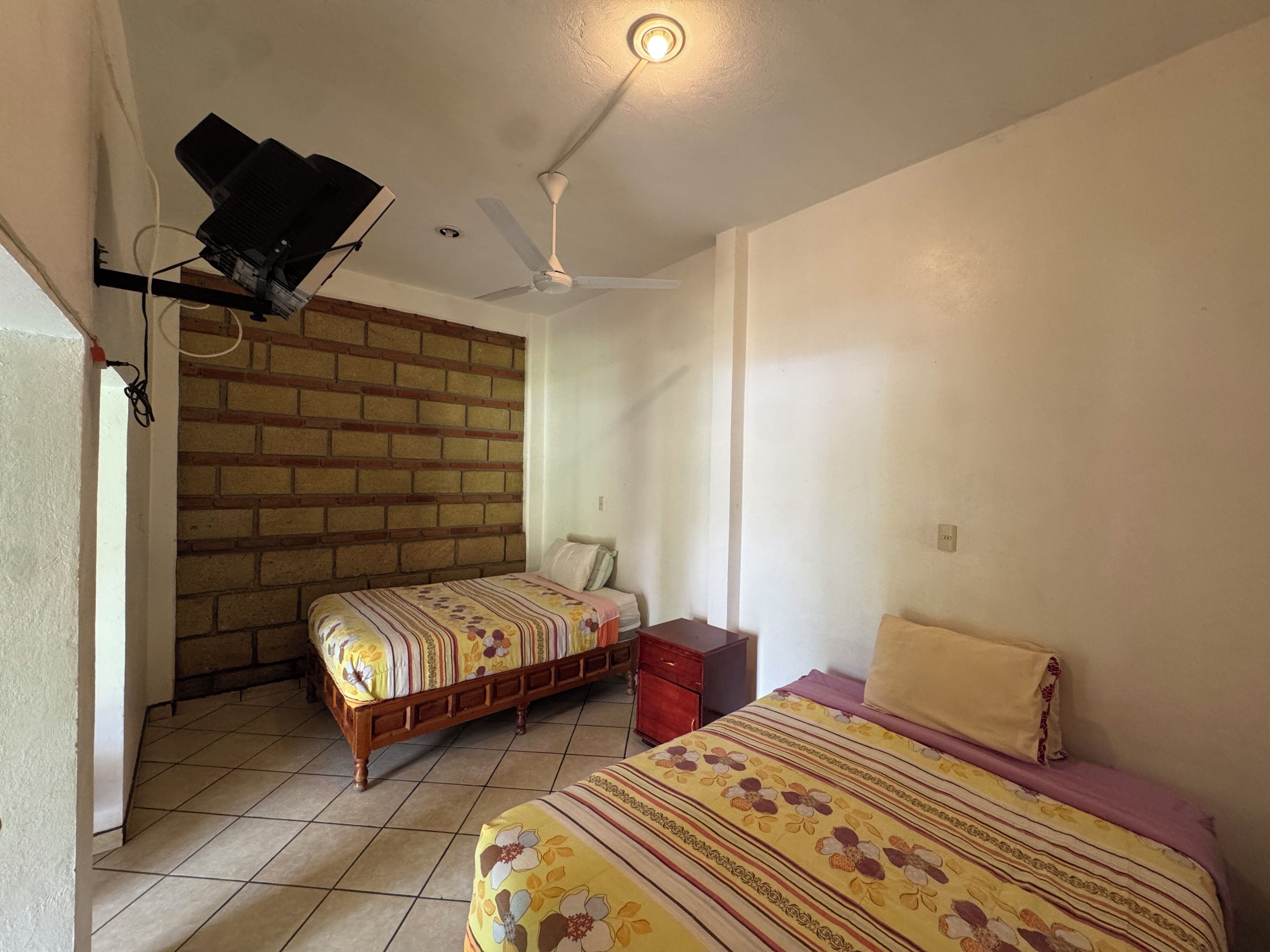 A bedroom with two beds, a ceiling fan, a brick wall, and a tiled floor.