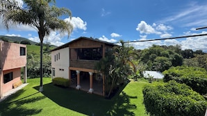 Una casa de dos pisos con un balcón, rodeada de exuberante vegetación y un cielo azul brillante.