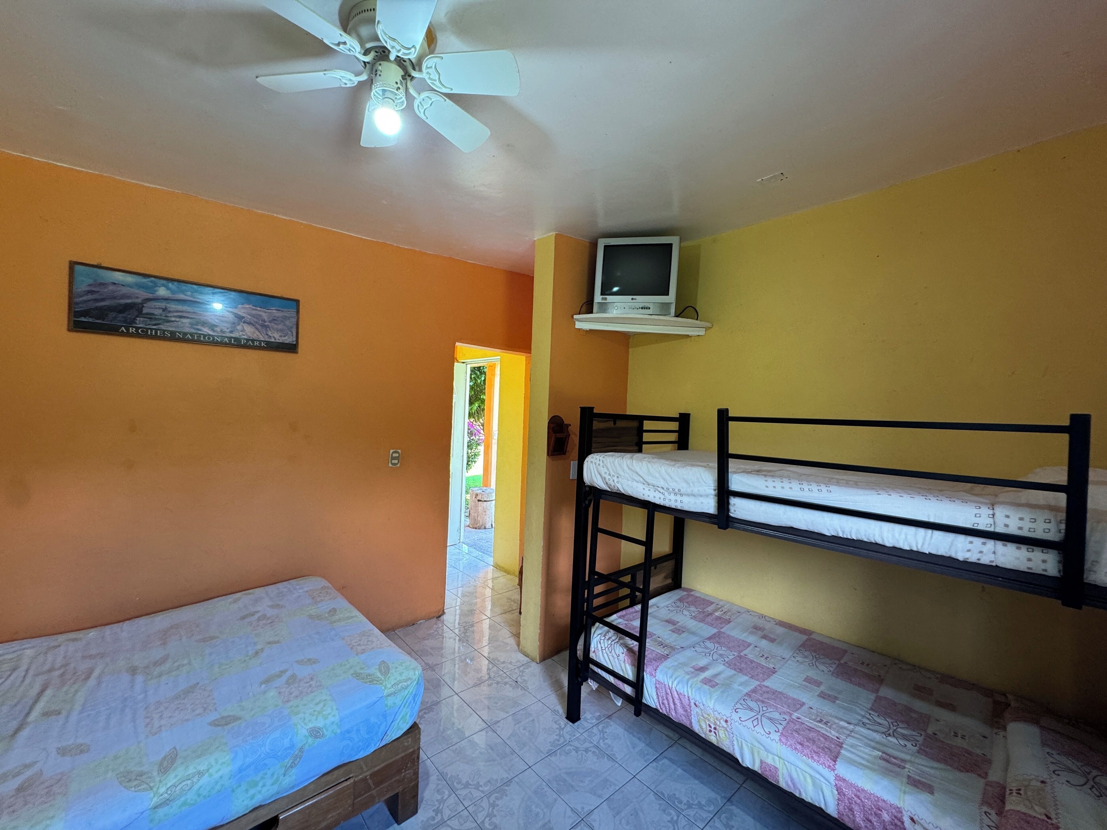 Una habitación con dos literas, un ventilador de techo, un televisor sobre una estantería y un cuadro en la pared.