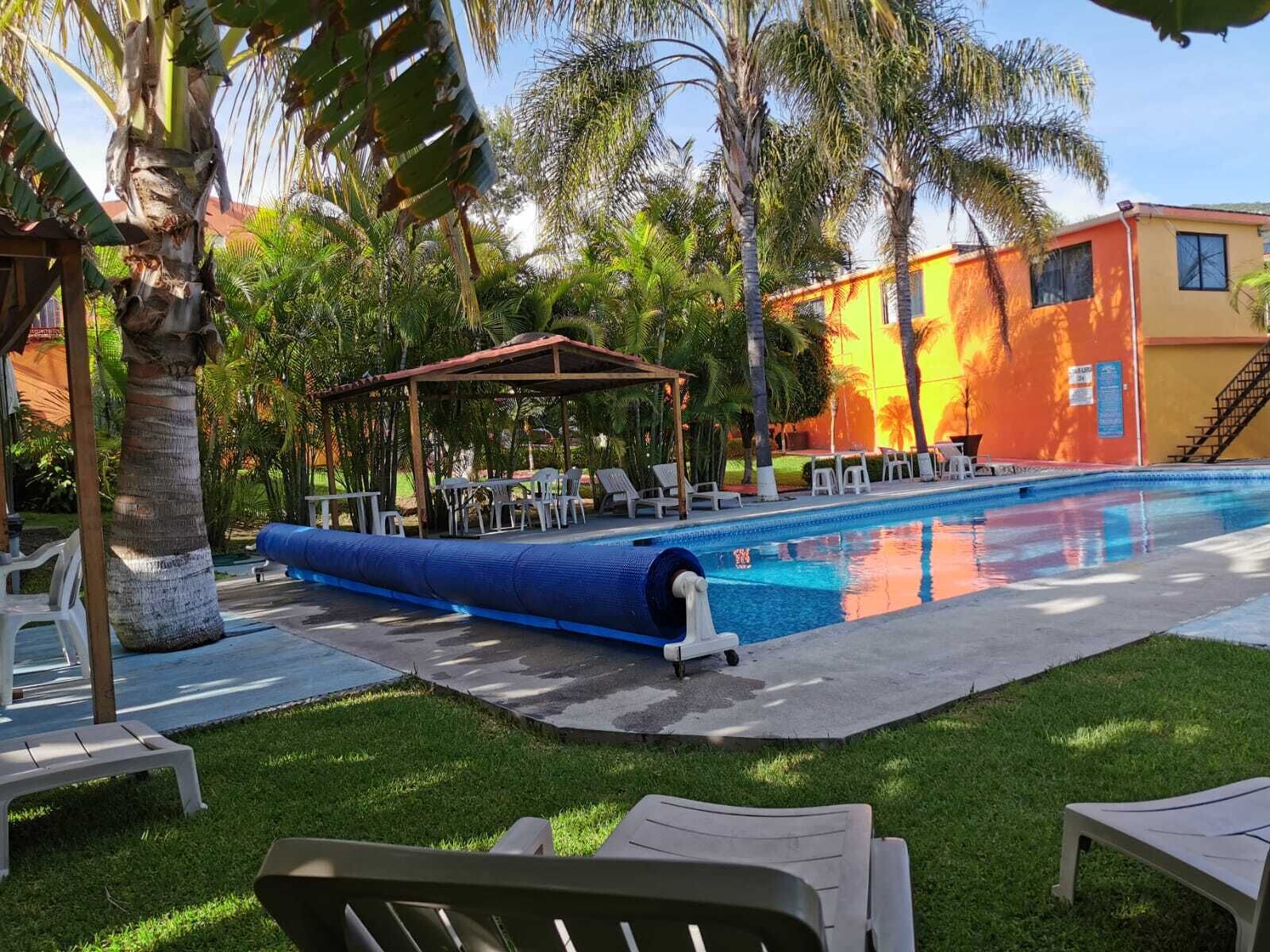 Un área de piscina con sillones de descanso, una zona de estar cubierta y un edificio de fachada amarilla.