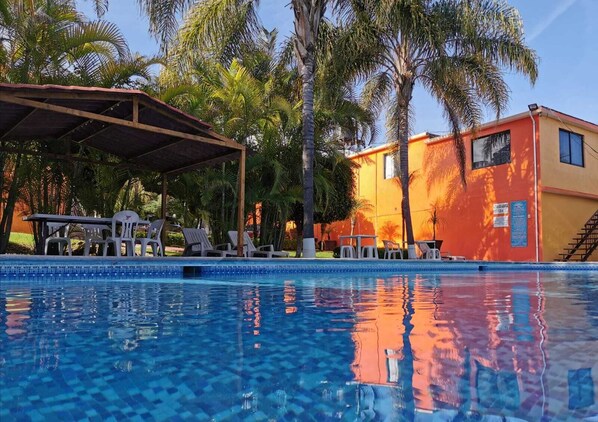 A swimming pool with lounge chairs and a covered area, surrounded by palm trees and a building with a red facade.