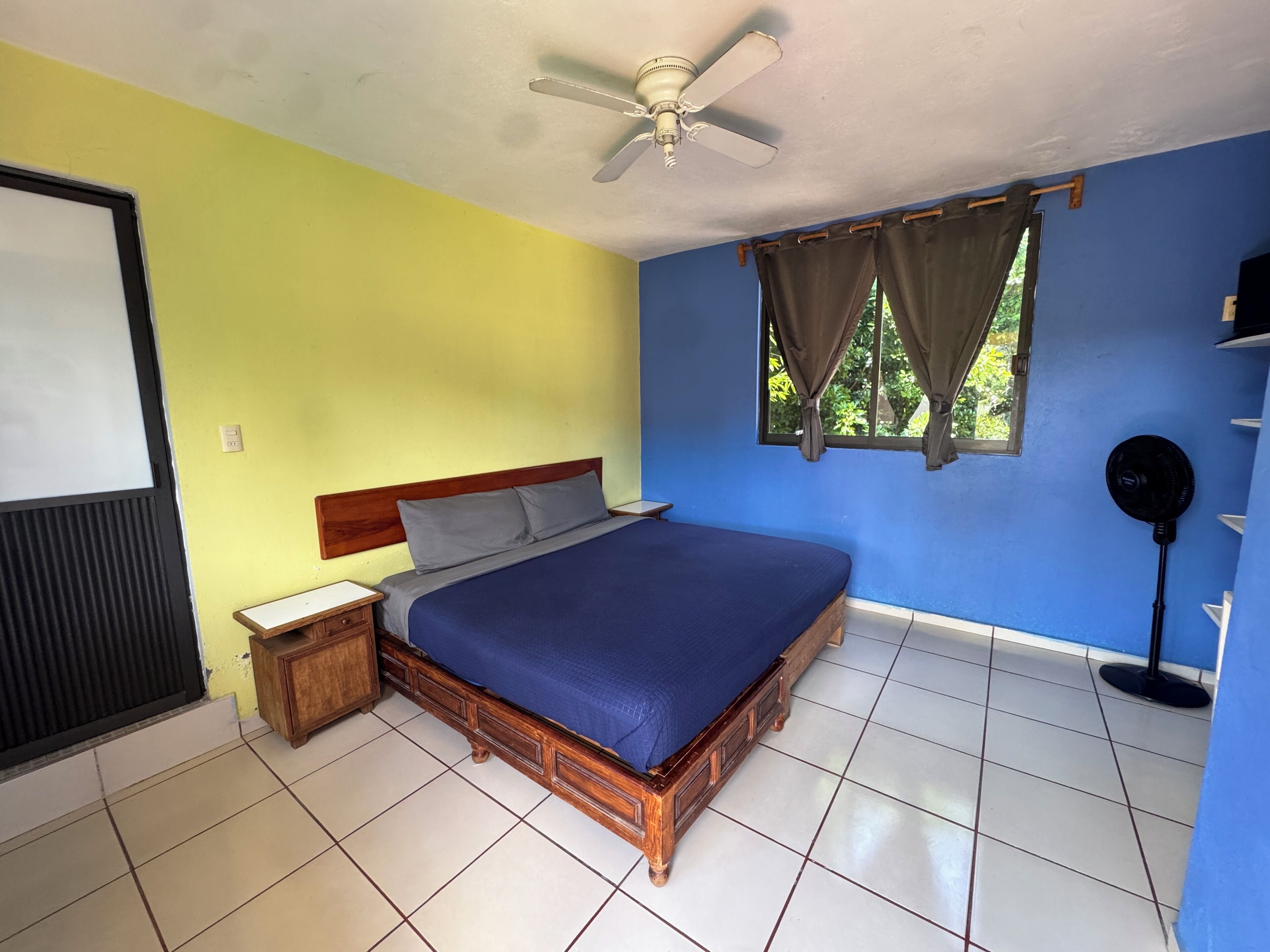 A bedroom with a wooden bed, a nightstand, a ceiling fan, a window with curtains, and a black speaker.