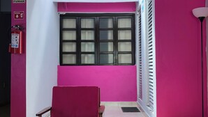 Um quarto com parede rosa, janela com persianas, uma cadeira vermelha, um extintor de incêndio e um controle remoto sobre uma mesa.