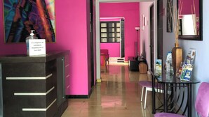 Um saguão de hotel com uma mesa de recepção, uma cadeira rosa e uma mesa de vidro com revistas.