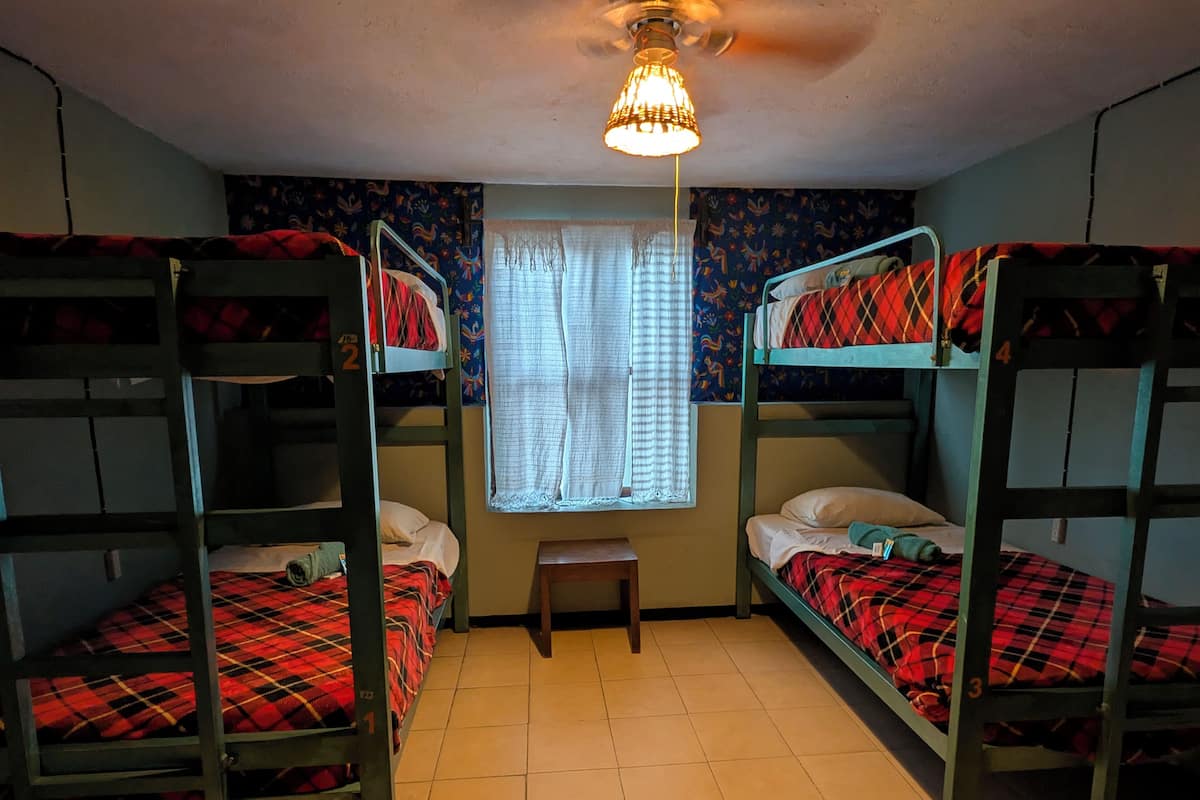 Um dormitório com beliches, uma mesinha e uma janela com cortinas.