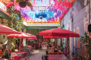 Un área de comedor al aire libre con mesas y sillas, sombrillas rojas y decoraciones colgantes.