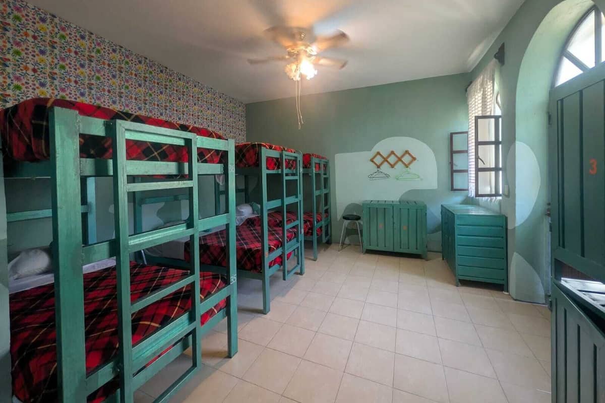Um dormitório com beliches, um ventilador de teto, uma escrivaninha pequena e uma janela.