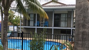 A pool area with a fenced pool, palm trees, and a building with barred windows in the background.