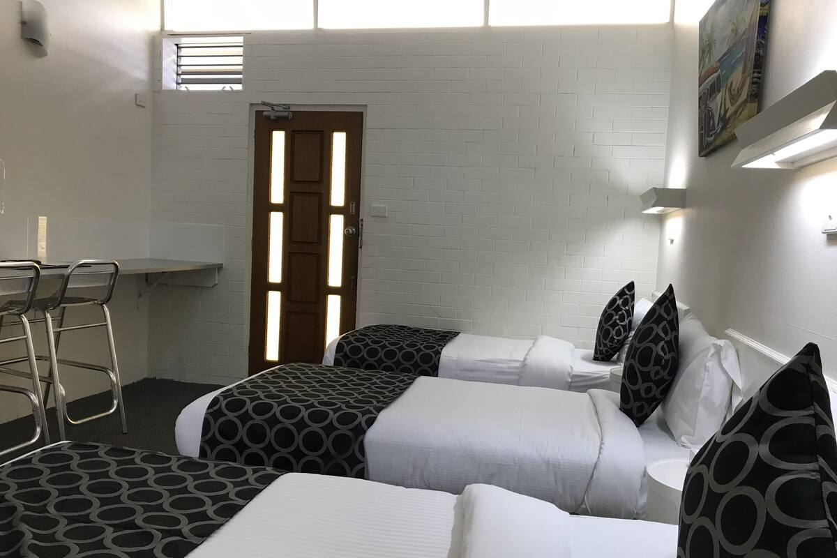 Quarto de hotel com duas camas, uma mesinha e uma cadeira. Há uma porta com janela e um quadro na parede.
