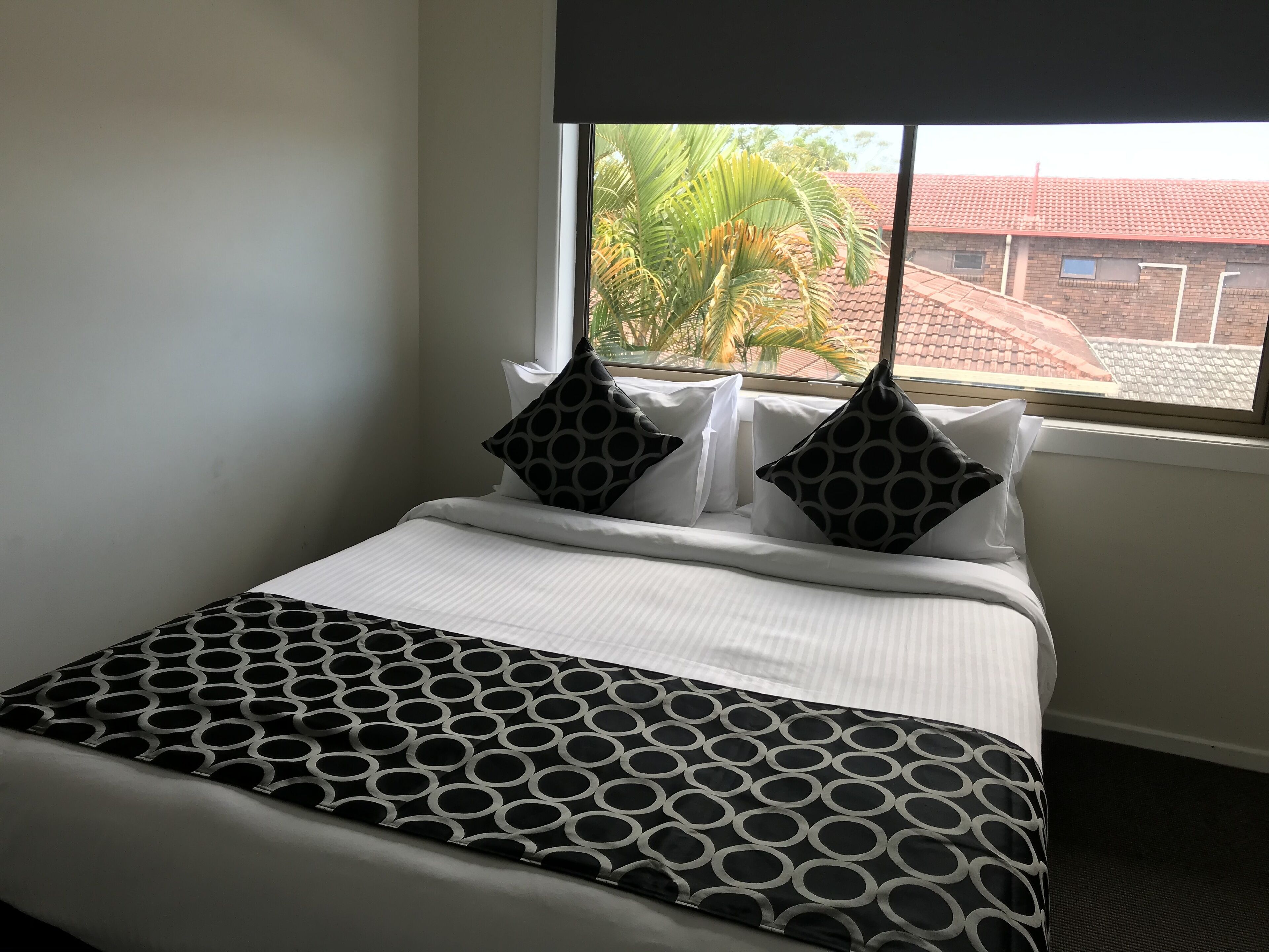 Un lit bien fait, avec une couvre-lit à motifs et deux oreillers, une fenêtre donnant sur un immeuble et des palmiers, et un mur sobre.