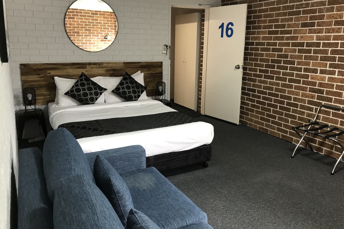 Quarto de hotel com cama, espelho redondo, parede com padrão de tijolos, porta com o número 16 e sofá azul.