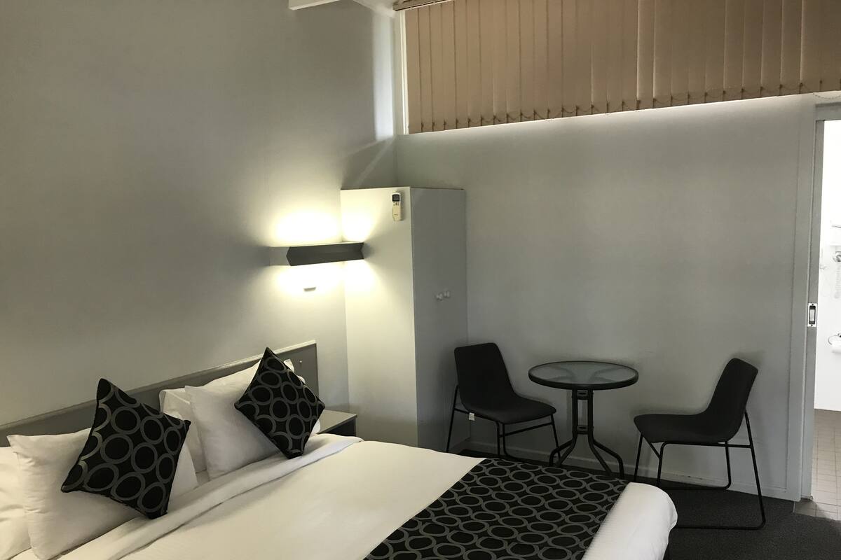 Quarto de hotel com uma cama, duas cadeiras, uma mesinha e ar condicionado fixado na parede.