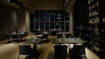 A modern restaurant with tables set for dining, a large window with a city view, and a wall of decorative vases.