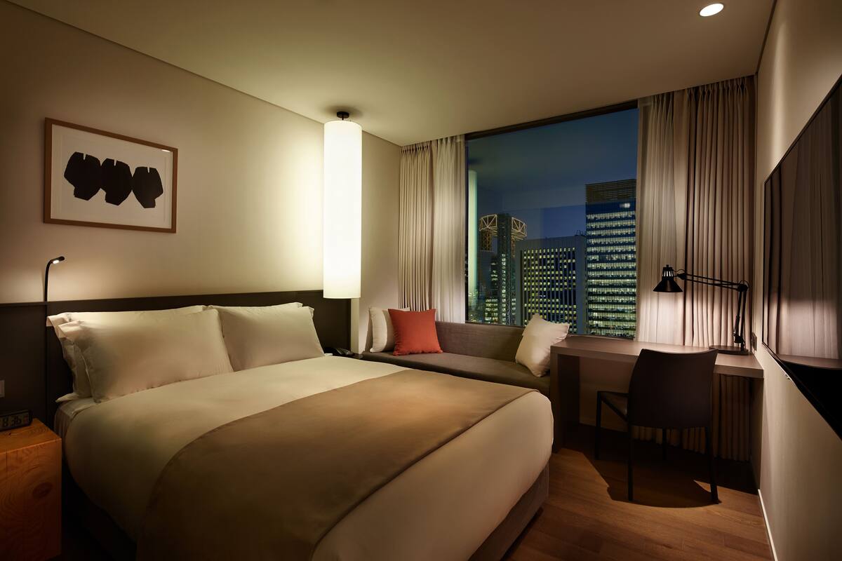 Une chambre d’hôtel avec un grand lit, un bureau et une vue sur le paysage urbain illuminé la nuit.
