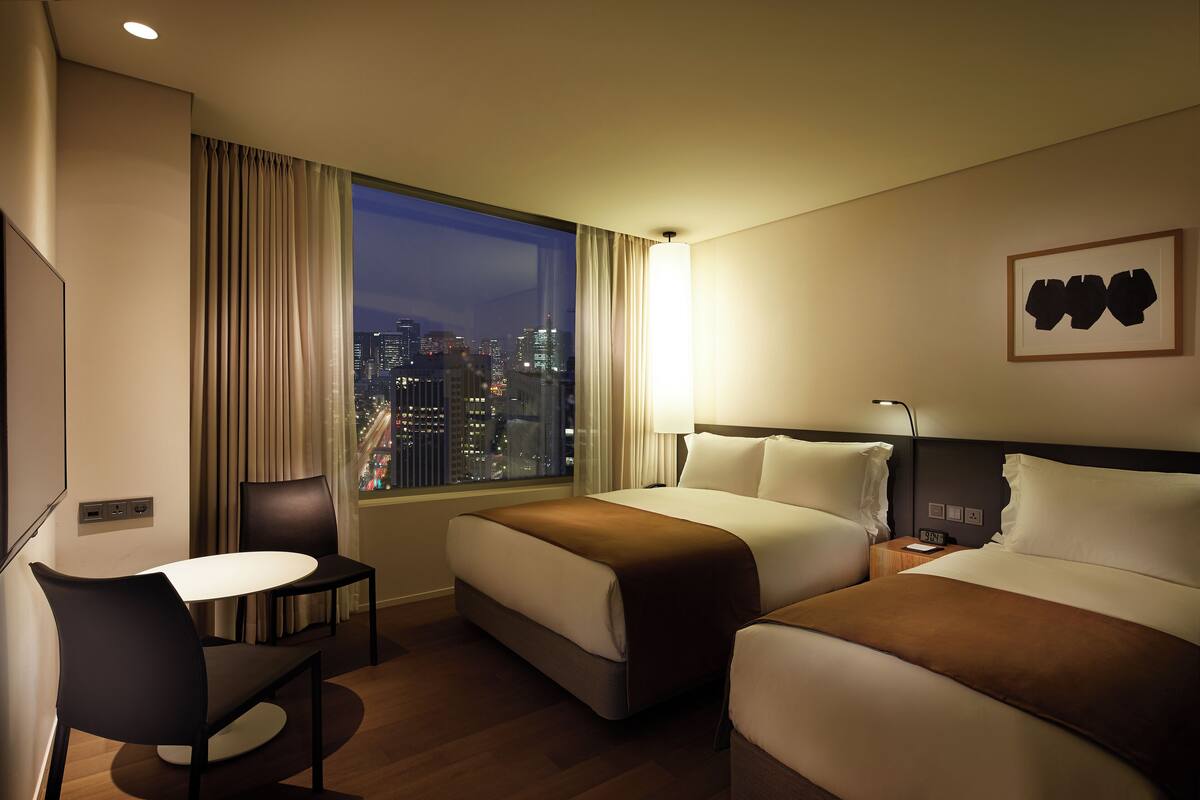 Une chambre d’hôtel avec deux lits, un bureau et une chaise. La fenêtre offre une vue sur la ville.