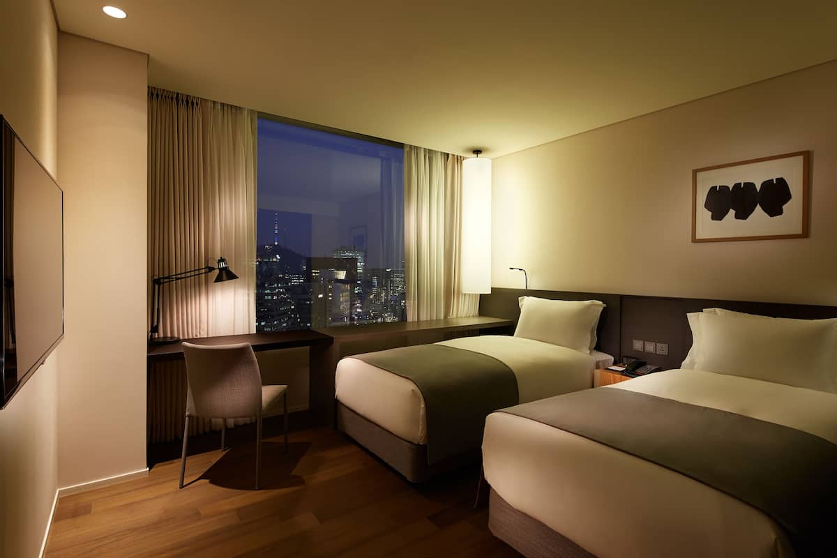 Une chambre d’hôtel avec deux lits, un bureau, une chaise, une vue sur la ville la nuit et une œuvre d’art encadrée au mur.