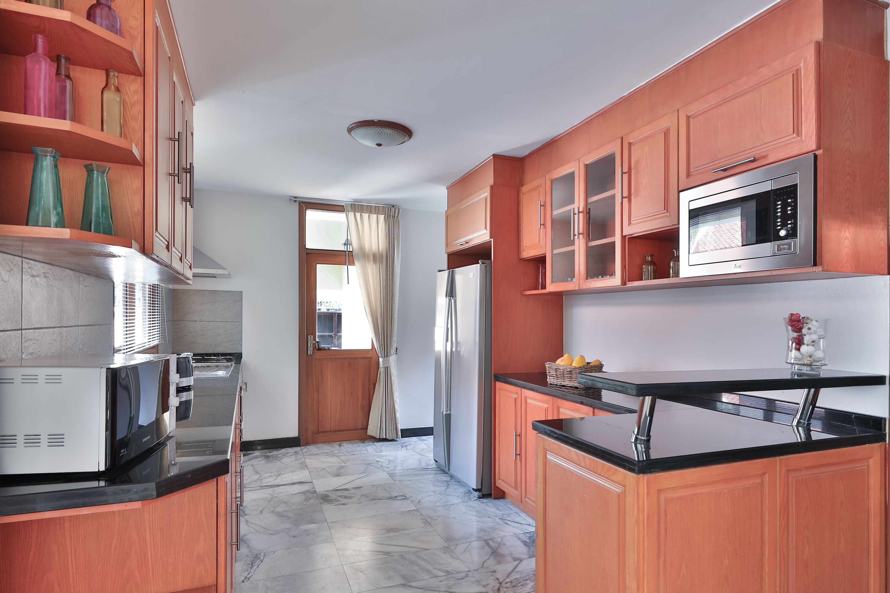 A kitchen with wooden cabinets, a refrigerator, a microwave, and a window with curtains.