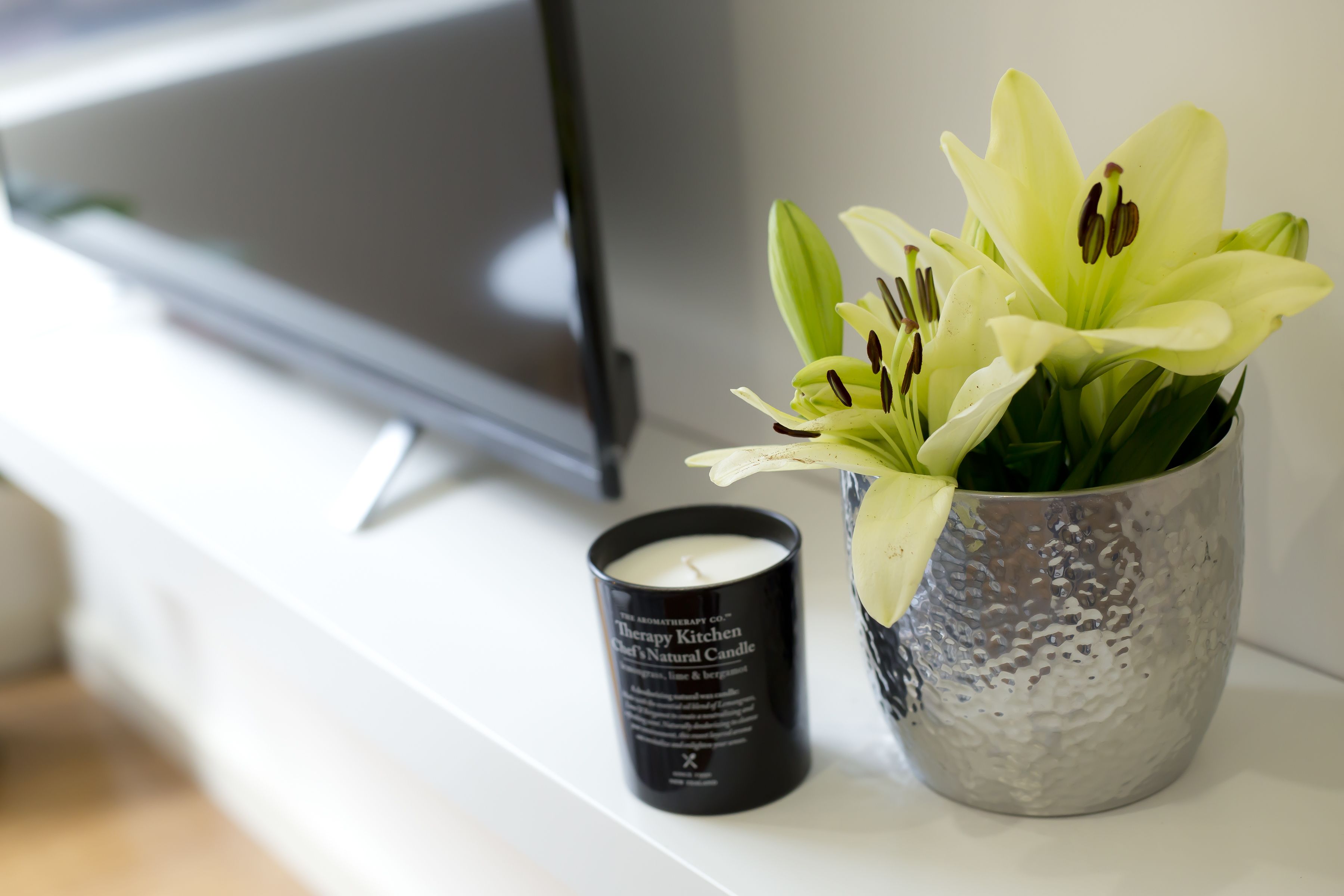 A white shelf with a black candle labeled "Therapy Kitchen" and a vase with white flowers.