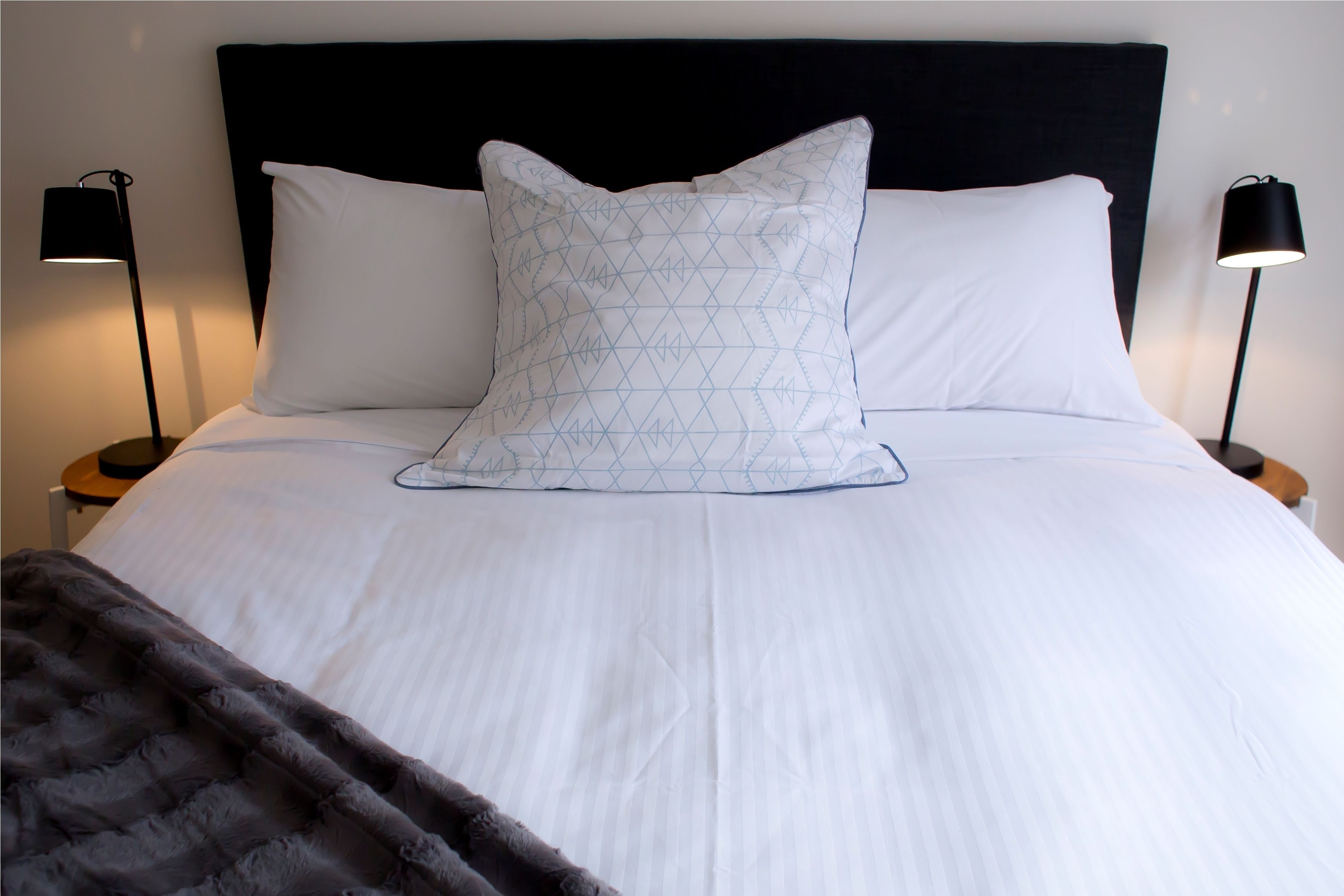 Una cama bien hecha con sábanas blancas y un cojín estampado, flanqueada por dos lámparas de noche negras.