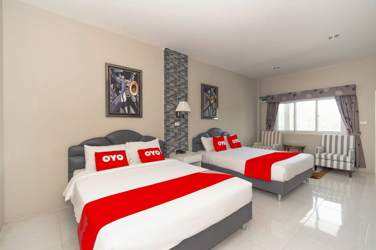 Een hotelkamer met twee bedden, elk met een rode en witte spreideken en kussens met 'OYO'-logo. Twee ingelijste kunstwerken hangen aan de muur, evenals een hoge, gegroefde kolom en een raam met gordijnen.