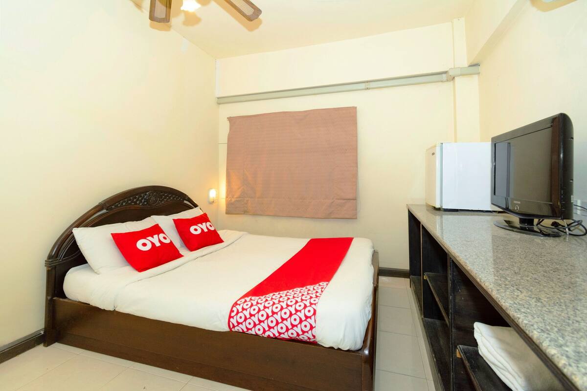 Een netjes opgemaakt bed met een wit en rode sprei, twee kussens met de tekst 'oyo', een televisie op een standaard en een bruin hoofdbord.