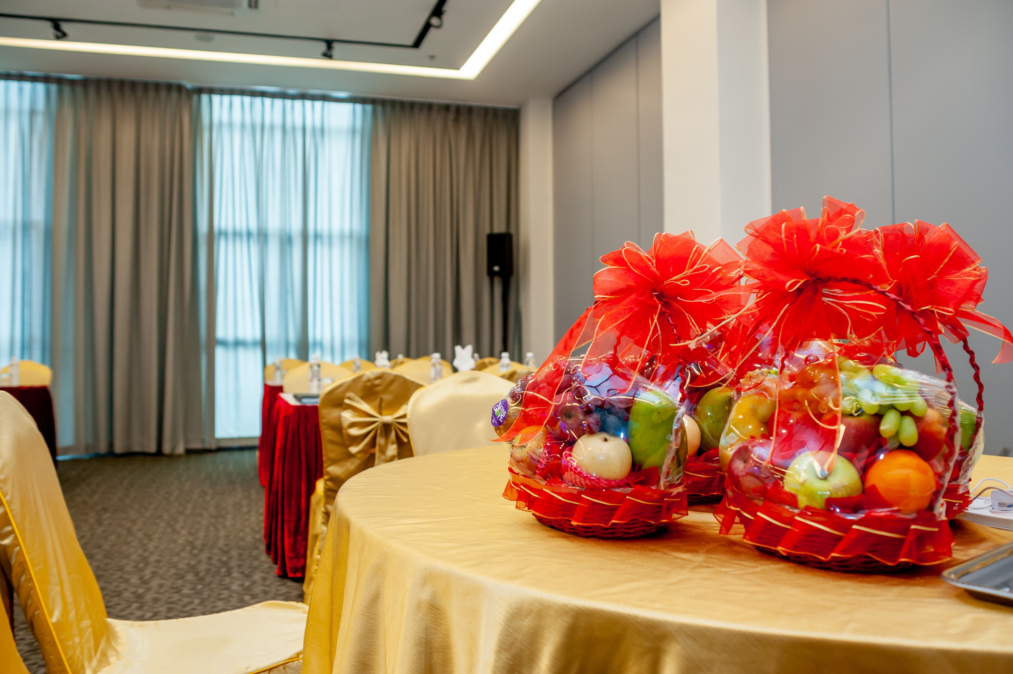 Habitación de hotel con una mesa redonda cubierta con mantel dorado, dos cestas de frutas adornadas con lazos rojos, y vistas de la habitación a través de amplias ventanas con cortinas.