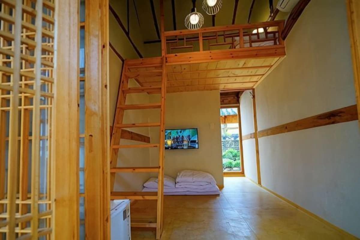 Une pièce avec un lit en mezzanine, une échelle, un petit canapé et un téléviseur fixé au mur.
