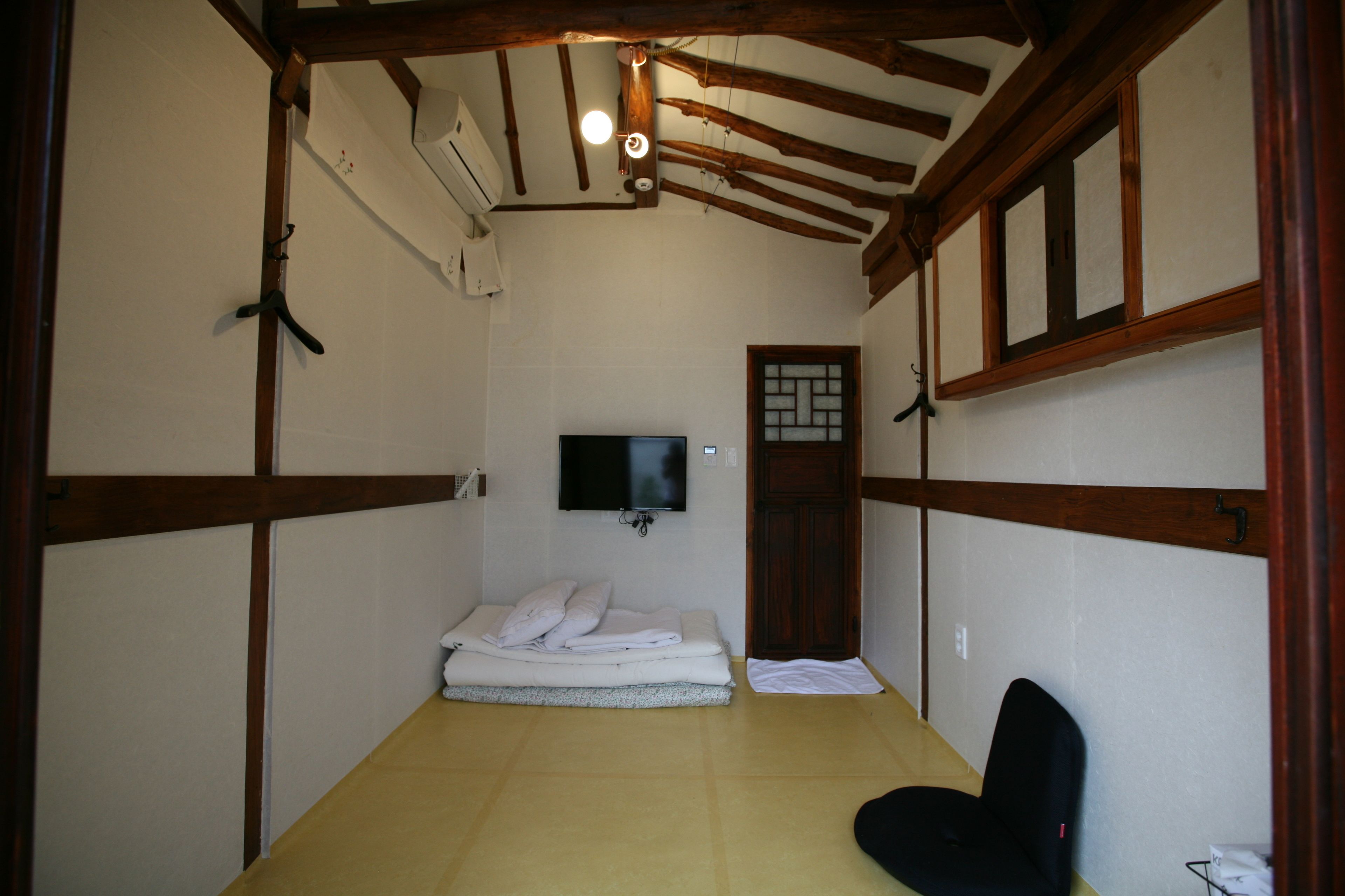 A small, traditional room with a tatami mat floor, a bed with a white blanket, a television on the wall, and a wooden door.