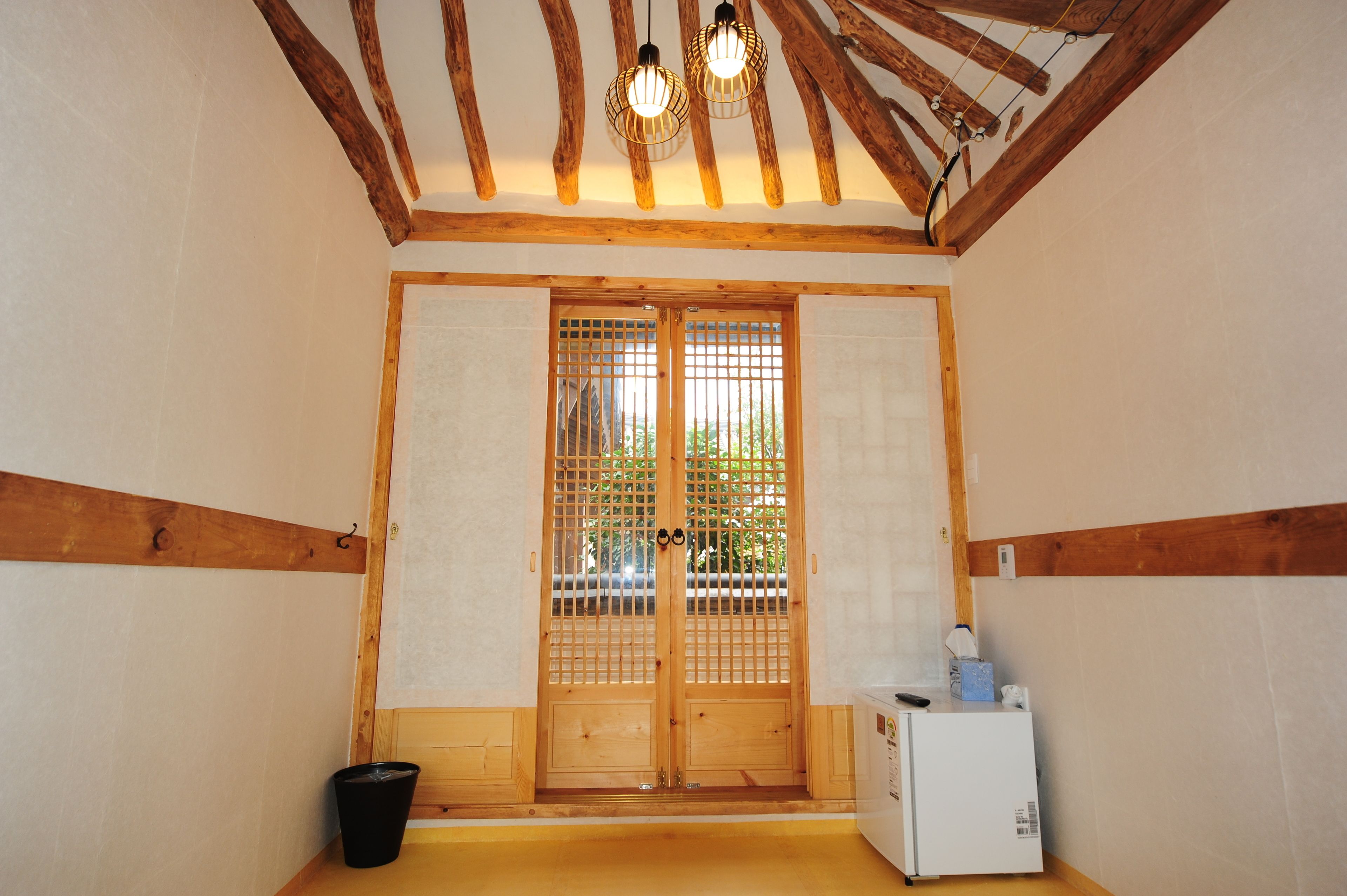 Una habitación tradicional japonesa con vigas de madera, un puertas correderas, un refrigerador pequeño y un cubo de basura negro.
