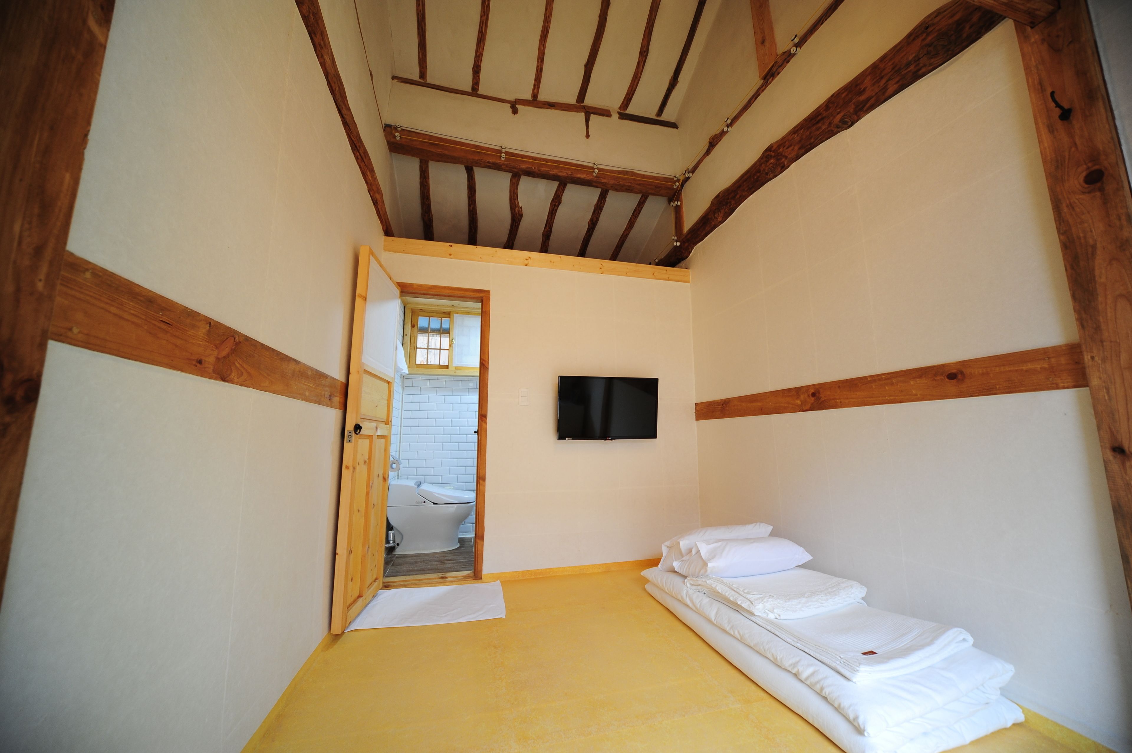 Une petite pièce étroite avec des poutres en bois au plafond, un lit recouvert de draps blancs, un téléviseur accroché au mur et une salle de bain visible à travers une porte ouverte.