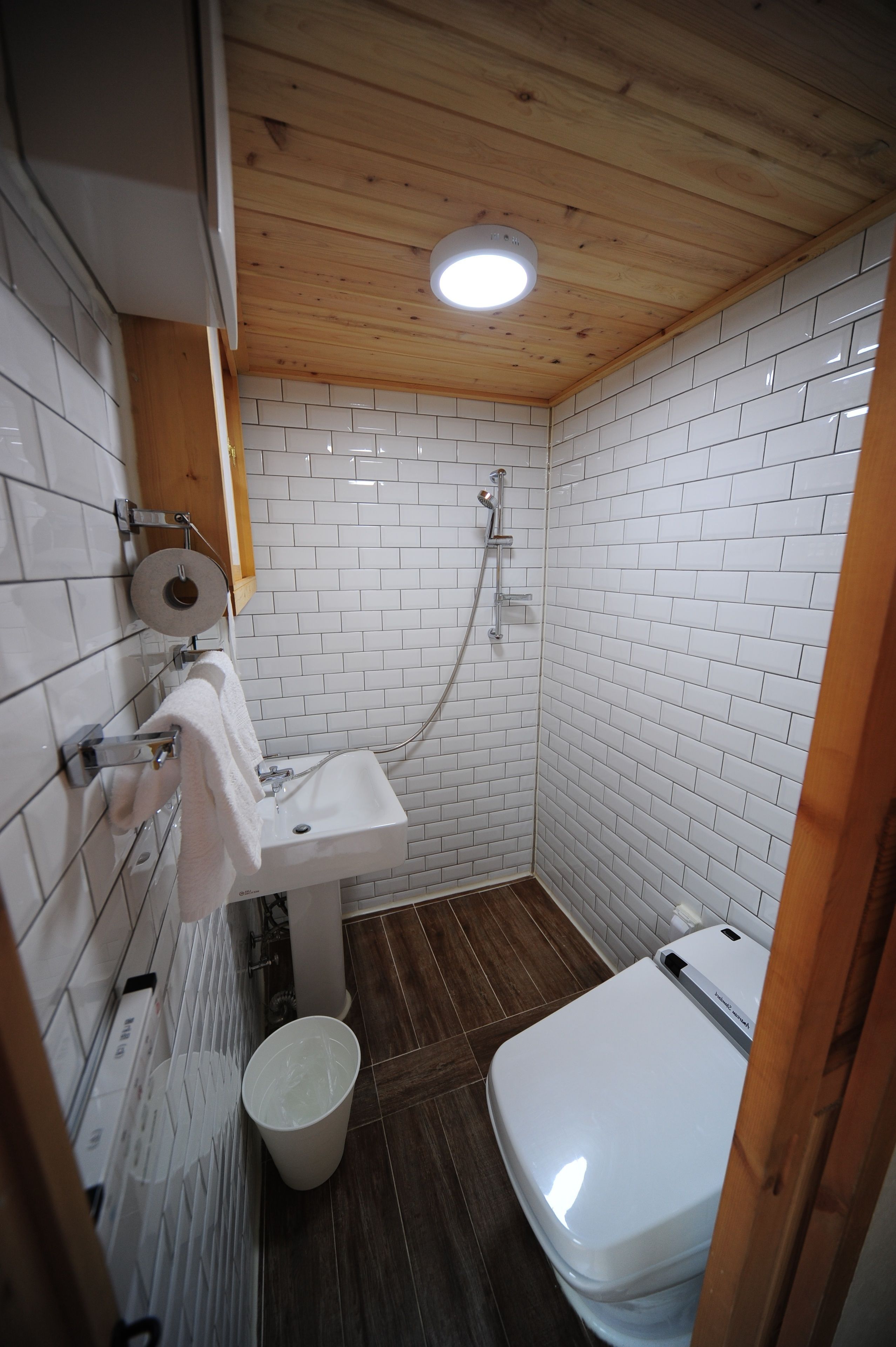 Une petite salle de bain compacte avec des murs recouverts de carreaux blancs, un plafond en bois, un lavabo blanc, des toilettes et une douche.