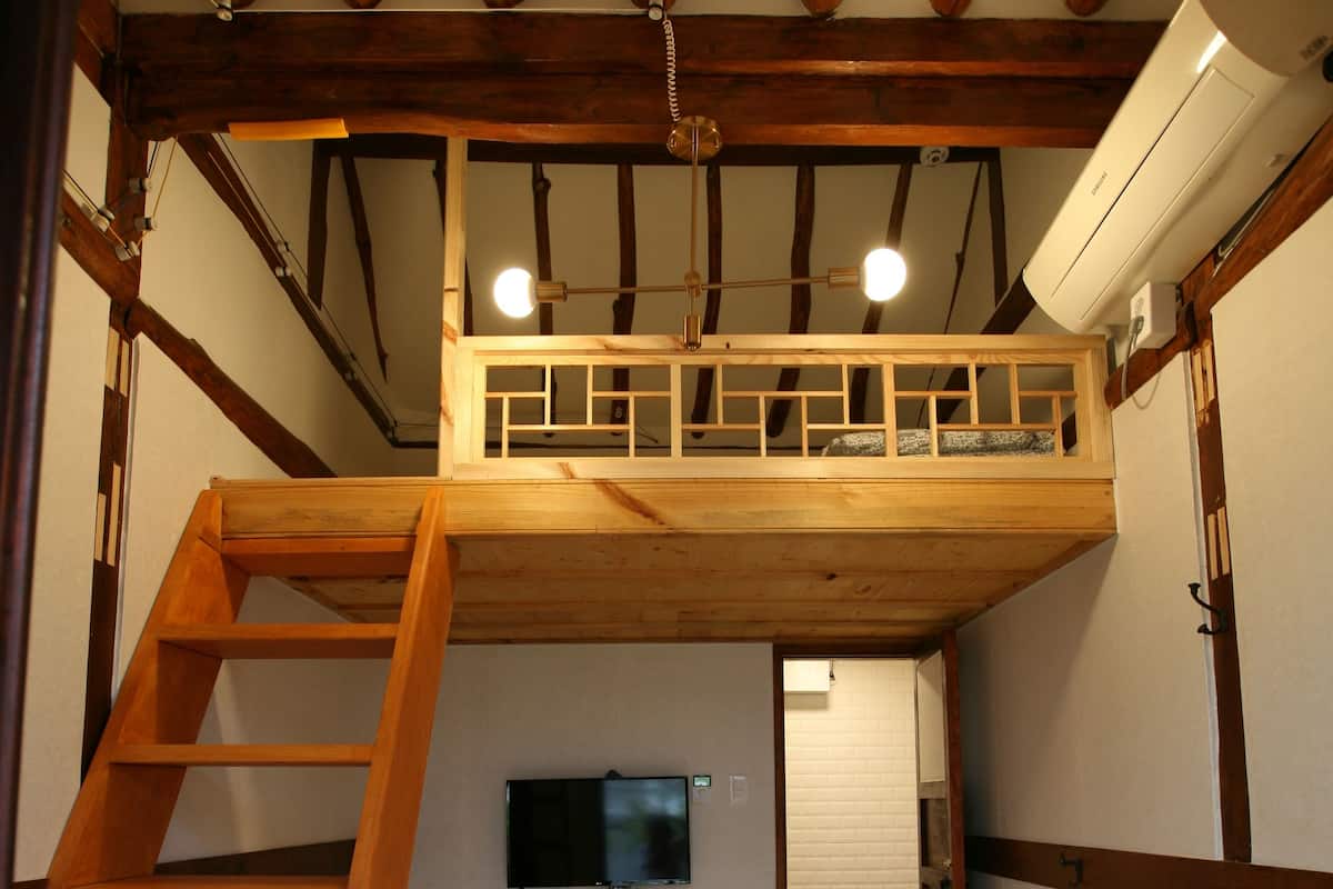 A compact room with a wooden bunk bed, a ladder, a flat-screen TV mounted on the wall, and a small entrance door.