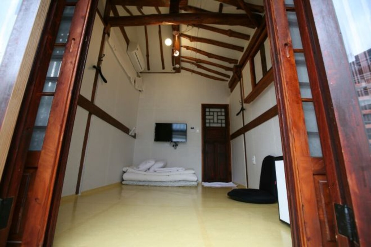 Une pièce étroite avec des poutres en bois, un lit recouvert de draps blancs, une chaise noire et une petite fenêtre.