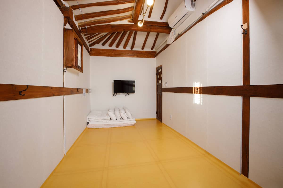 A room with a yellow floor, wooden beams, a flat-screen TV mounted on the wall, and a stack of white towels against the wall.