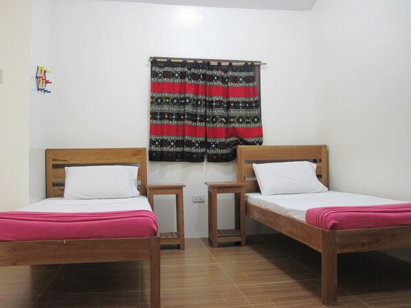 A room with two wooden beds, each with a pink bedspread and white pillows. There is a small wooden nightstand between the beds. A colorful curtain hangs on the window behind the beds.
