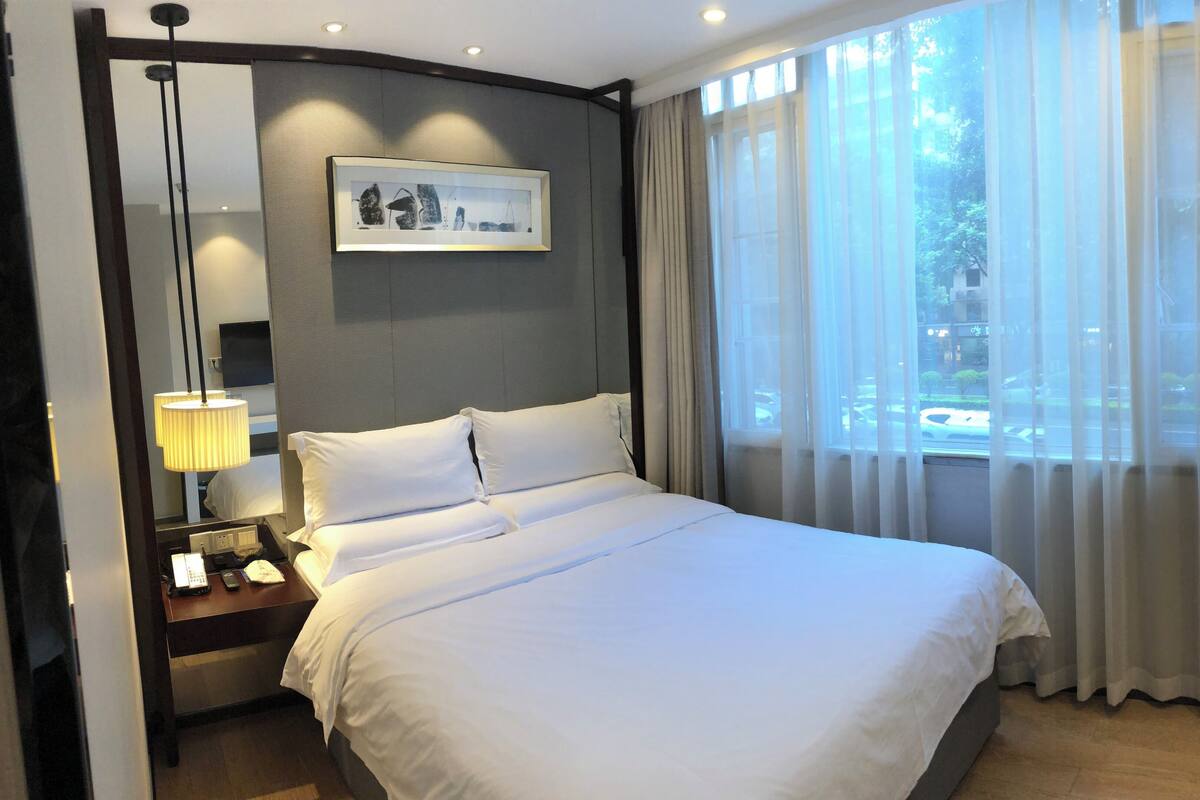 A hotel room with a bed, bedside table, lamp, and a window with curtains.