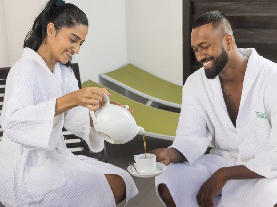 Two individuals in white robes seated on a bench, one pouring a beverage into a cup.