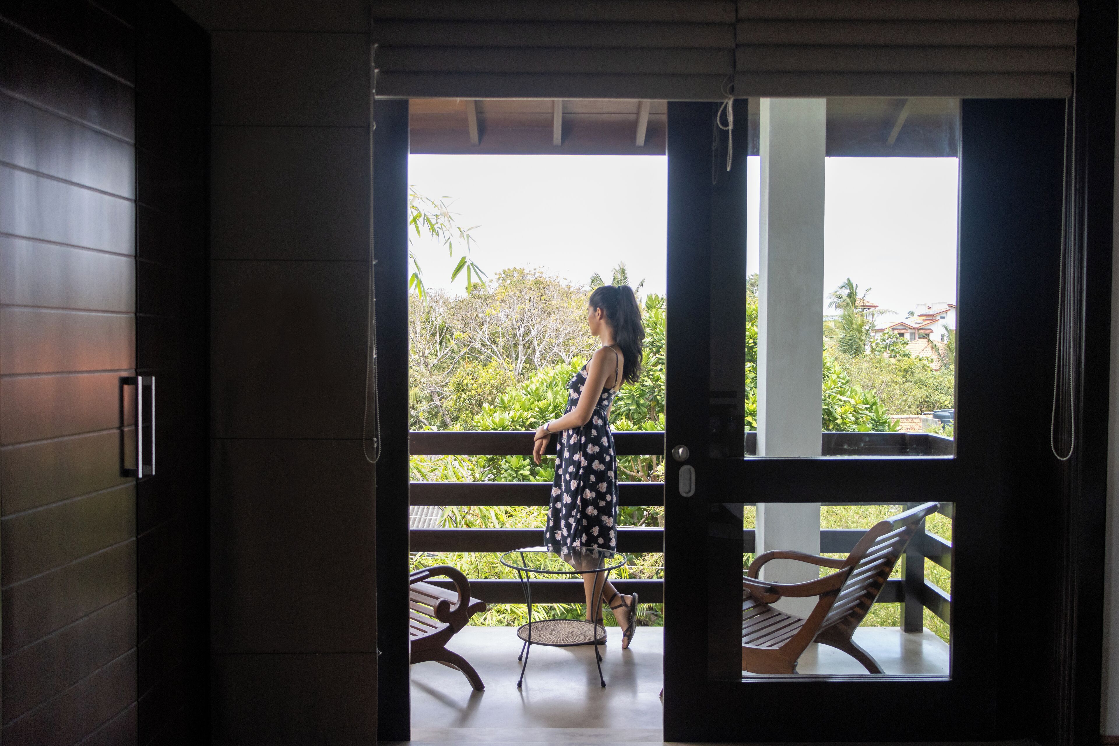 A woman in a dress standing on a balcony with a view of greenery and buildings.