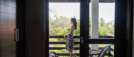 A woman in a dress standing on a balcony with a view of greenery and buildings.