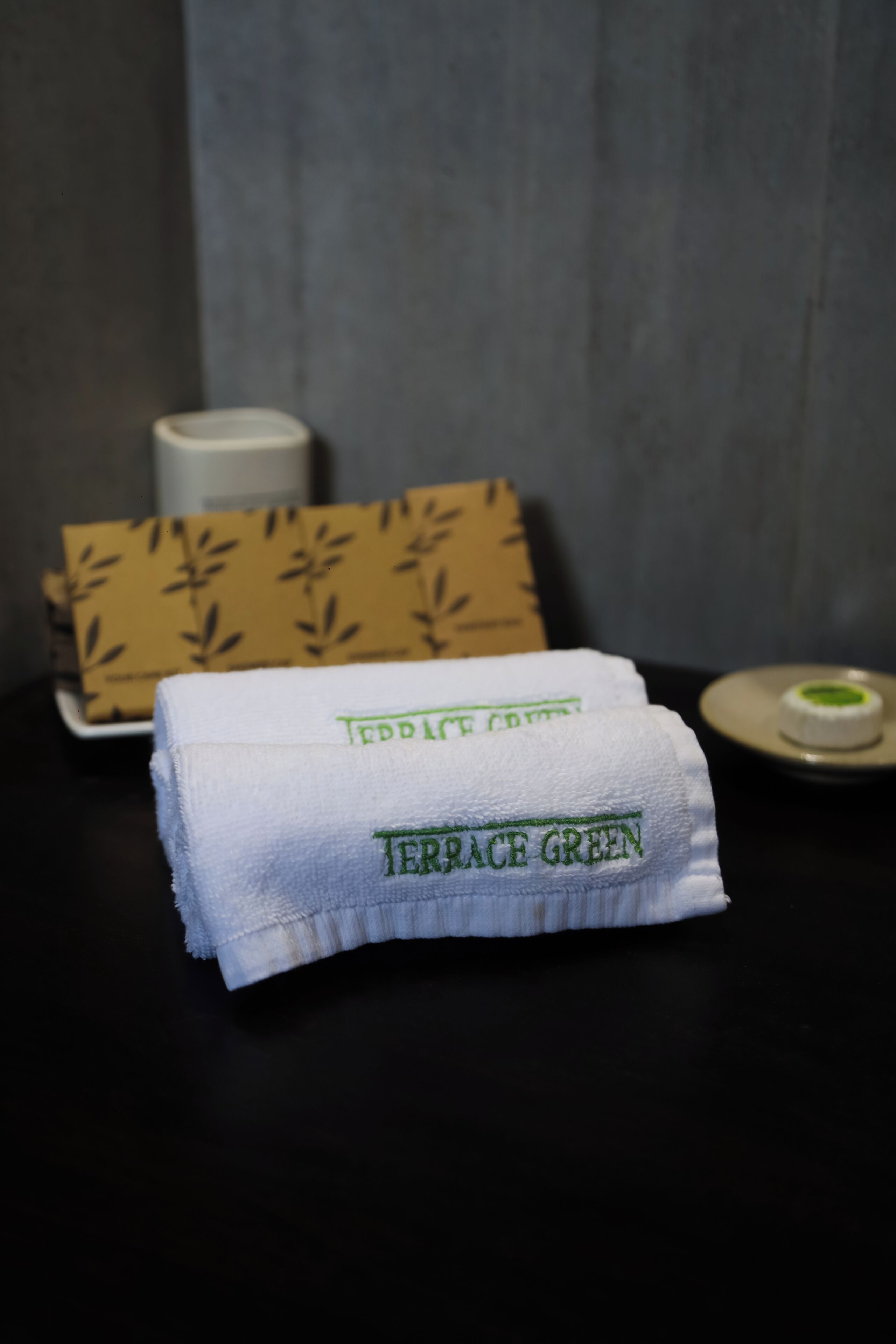 A neatly folded towel with "Terrace Green" embroidered on it, placed on a dark surface with a patterned box and a small container in the background.