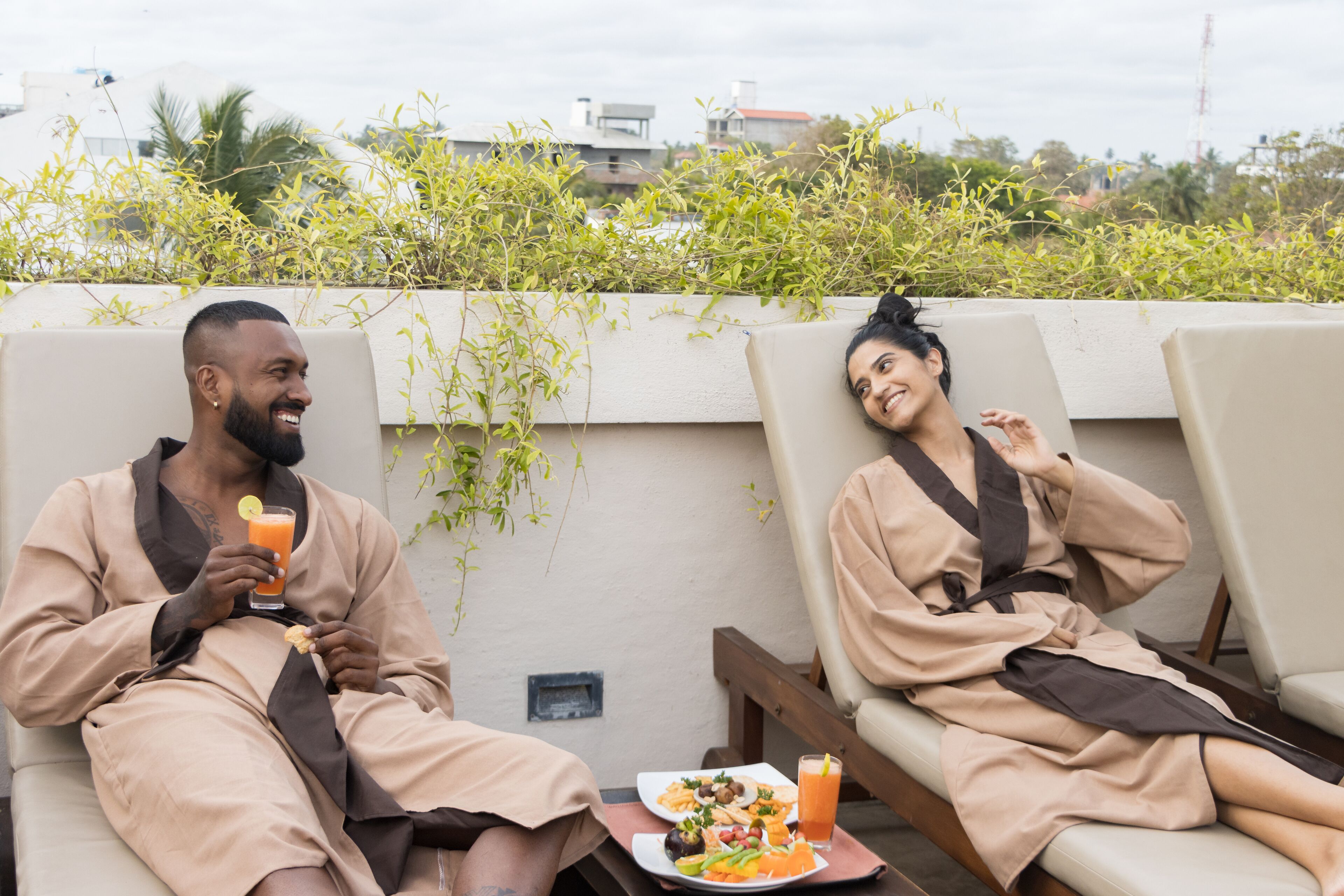 Two people in beige robes sitting on lounge chairs with a table of food and drinks between them.