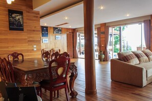 A spacious room with a wooden dining table, ornate chairs, a sofa, and large windows offering a view of the outdoors.