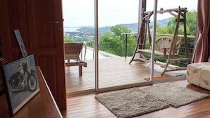 A room with a wooden floor, a sofa, a wooden swing set, and a view of a landscape with hills and a body of water.